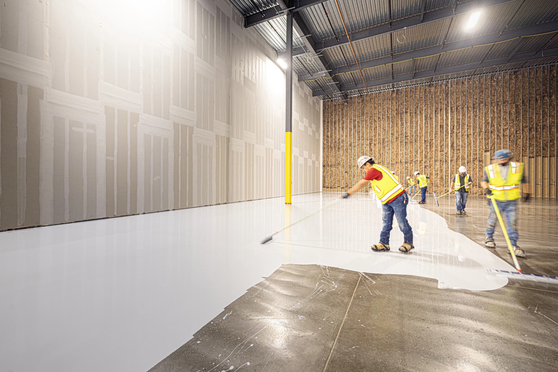 Construction crew using squeegees to apply a smooth white epoxy floor coating in a large warehouse.