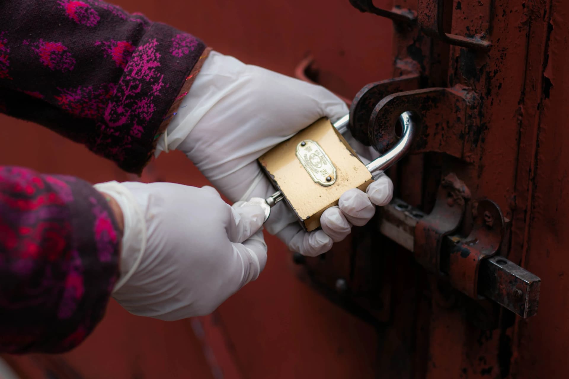 Hands wearing white rubber gloves using a key to unlock a brass padlock on a red metal gate.