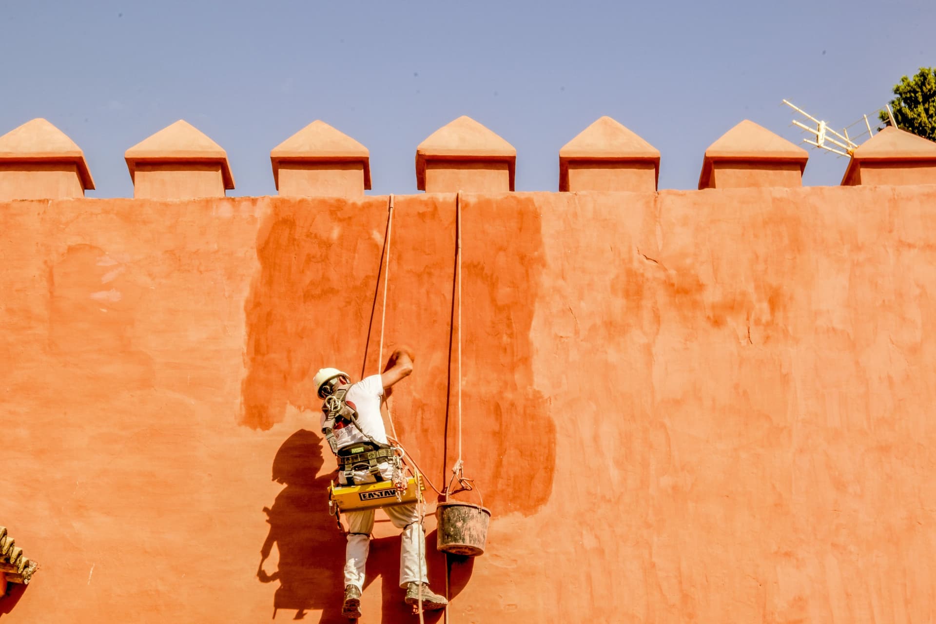 Painter suspended on ropes applying orange paint to a high exterior wall.