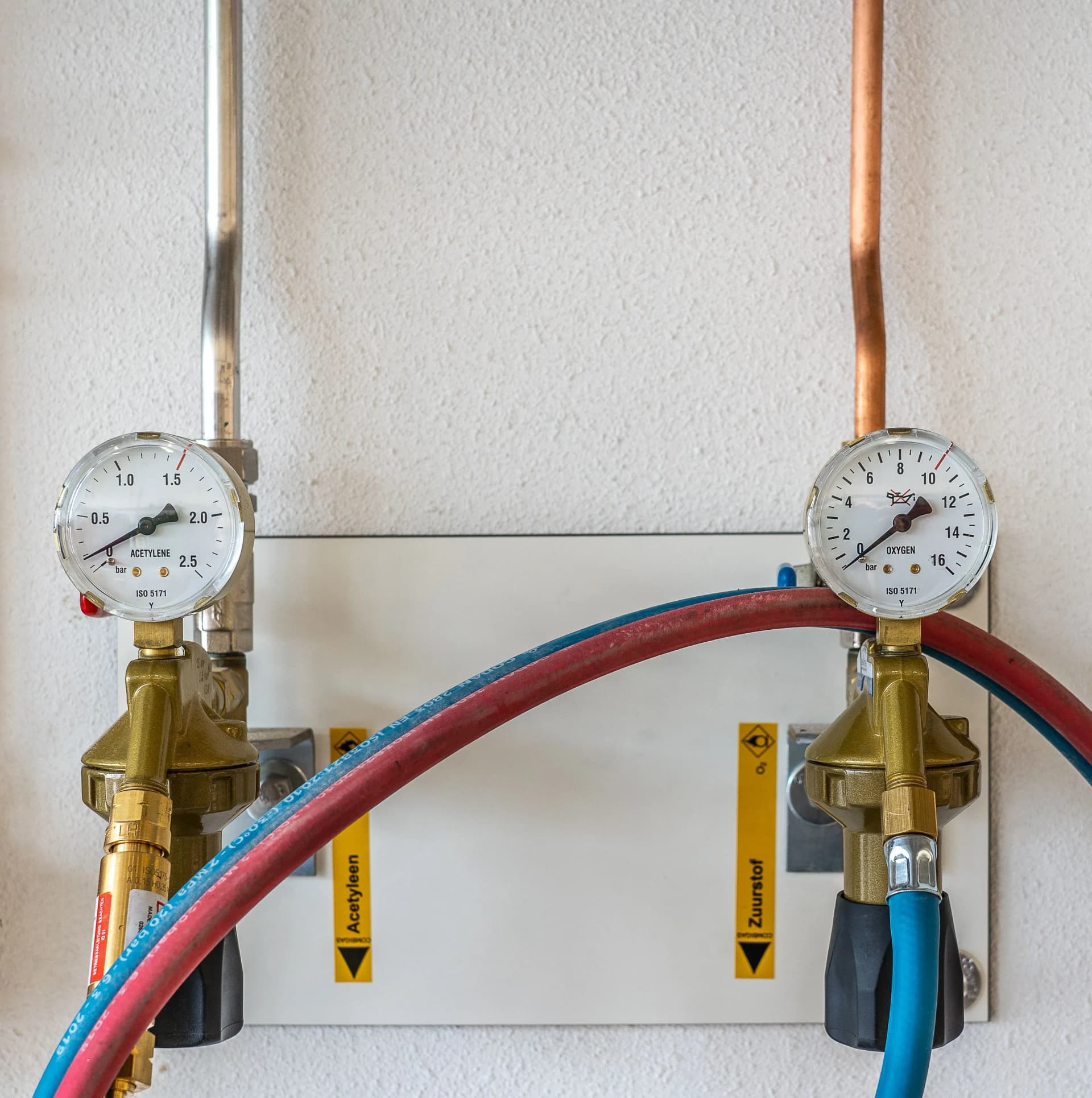 Close-up of industrial gas pressure gauges connected with hoses against a wall.