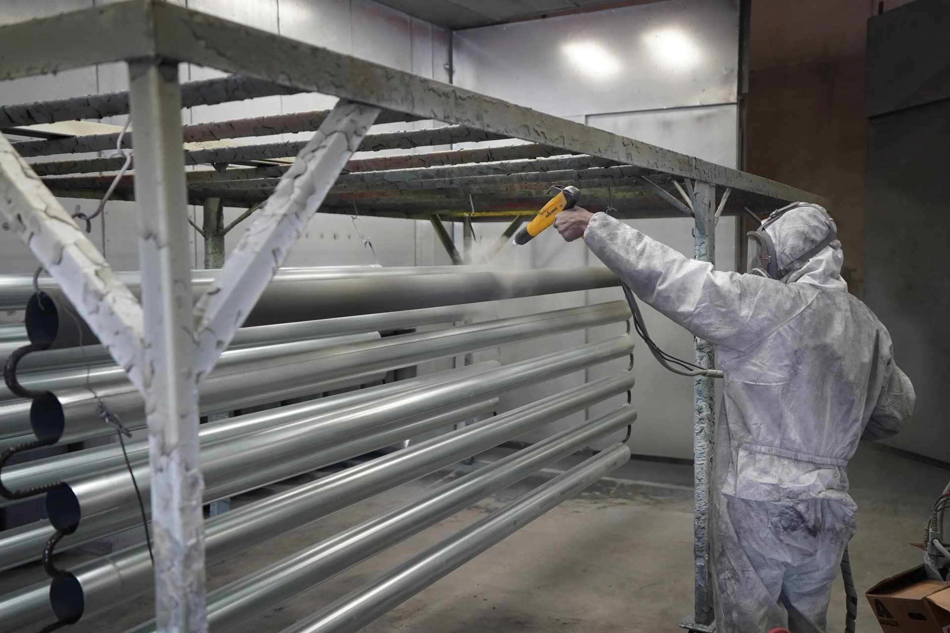 Worker in protective coveralls using a powder coating gun to spray metal pipes on a rack.
