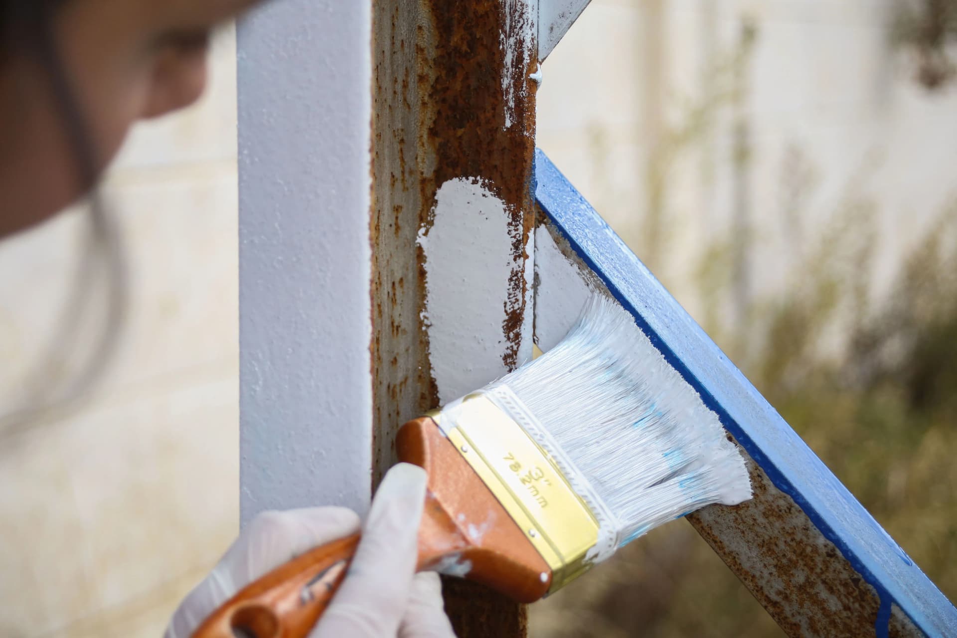 Close-up of a worker using a paintbrush to apply white paint over a rusted metal beam.