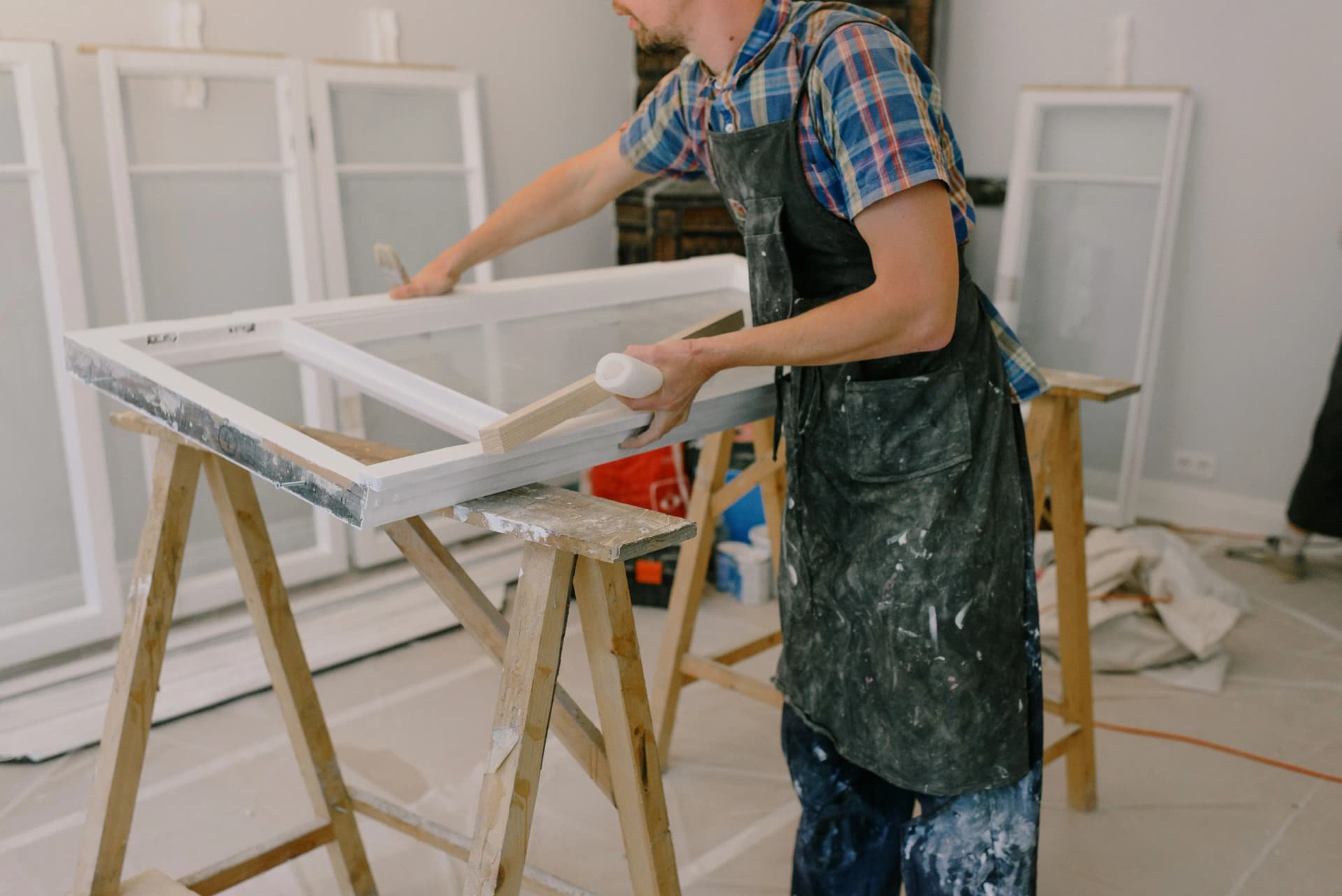 Craftsman applying white paint to a wooden window frame on saw horses.