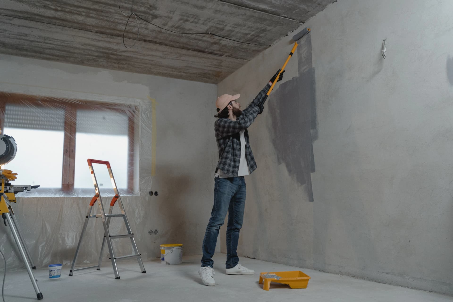 Man using a long paint roller to apply grey paint to an unfinished concrete wall.