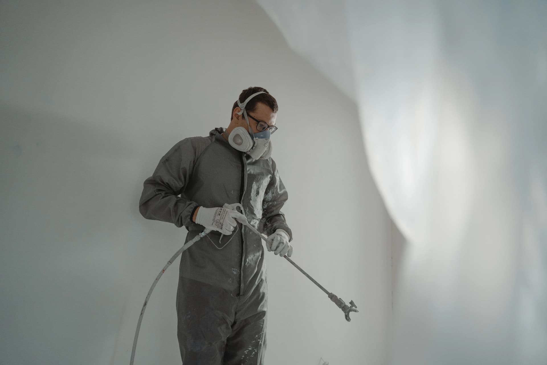 Worker wearing a respirator mask and protective suit using an airless paint sprayer on a wall.