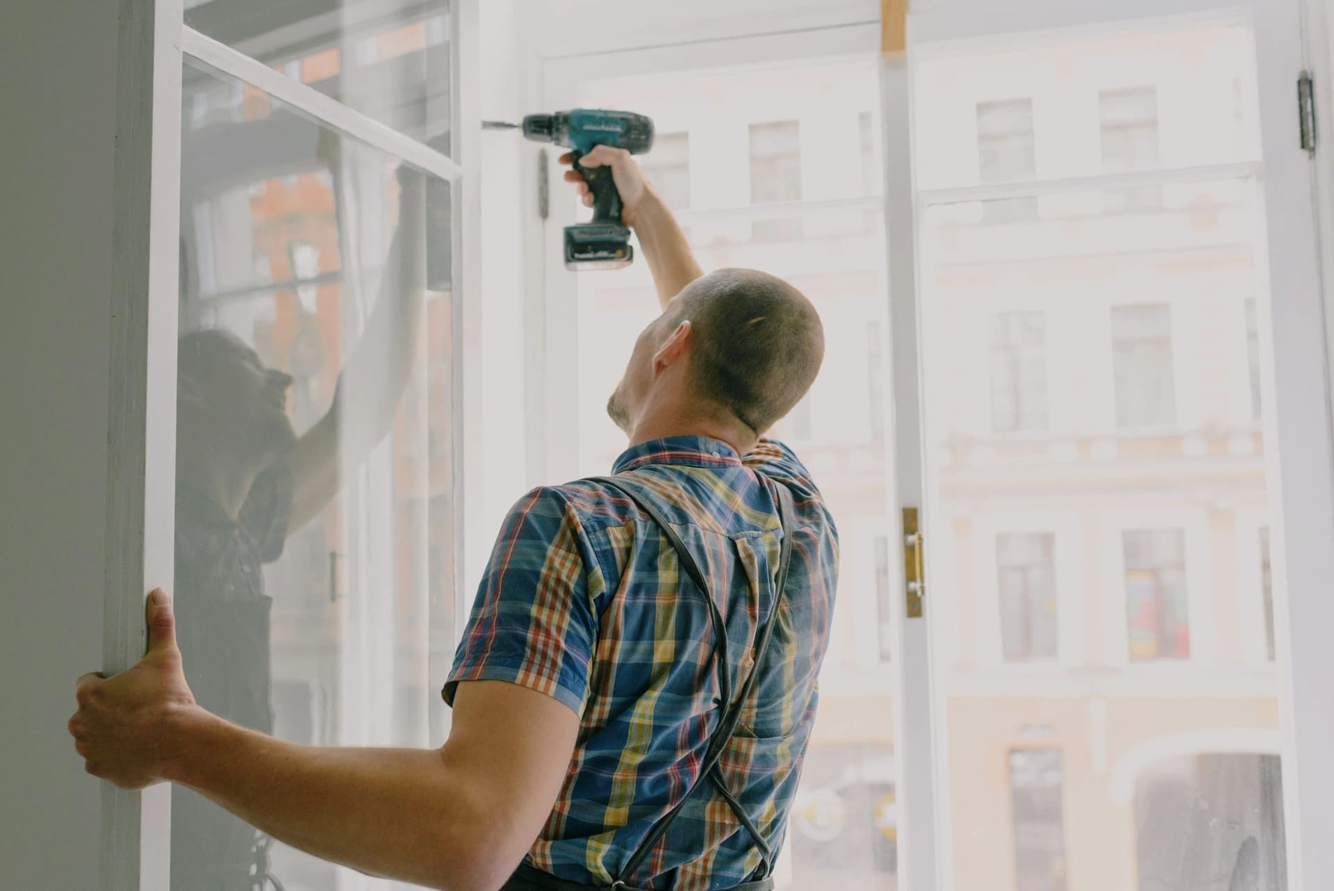 Carpenter using an electric drill on top of a tall white window frame.